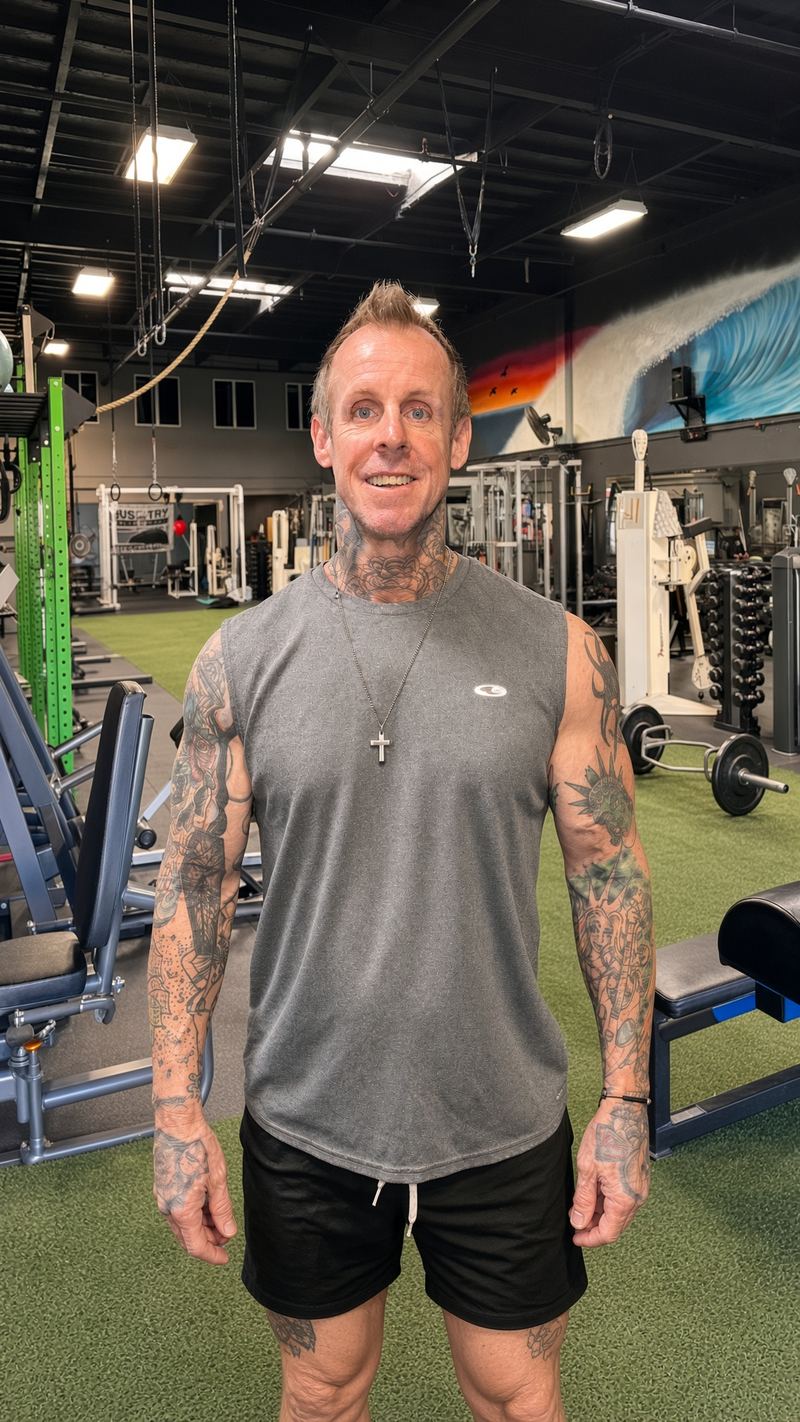 Mark Thomas, owner and trainer, smiling in a grey sleeveless tank with a cross necklace, standing on the turf in front of the gym equipment.